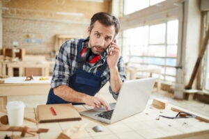 Artisan menuisier en salopette bleue et chemise à carreaux, assis à son établi dans son atelier. Il a un casque antibruit autour du cou, un téléphone à l’oreille et tape sur un ordinateur portable ouvert, regardant fixement la caméra.