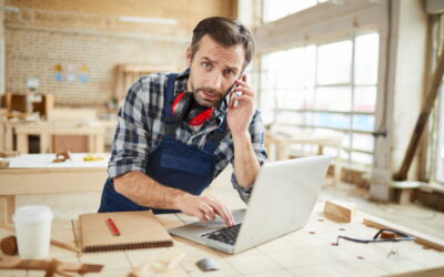 Artisan menuisier en salopette bleue et chemise à carreaux, assis à son établi dans son atelier. Il a un casque antibruit autour du cou, un téléphone à l’oreille et tape sur un ordinateur portable ouvert, regardant fixement la caméra.