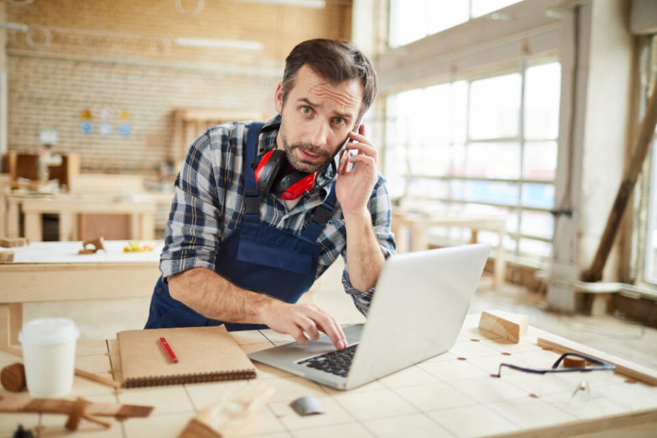 Artisan menuisier en salopette bleue et chemise à carreaux, assis à son établi dans son atelier. Il a un casque antibruit autour du cou, un téléphone à l’oreille et tape sur un ordinateur portable ouvert, regardant fixement la caméra.