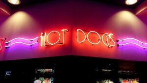 Panneau néon "HOT DOGS" en rouge et violet sur un mur au-dessus d'un comptoir avec écrans