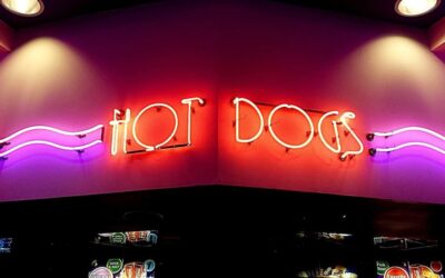 Panneau néon "HOT DOGS" en rouge et violet sur un mur au-dessus d'un comptoir avec écrans